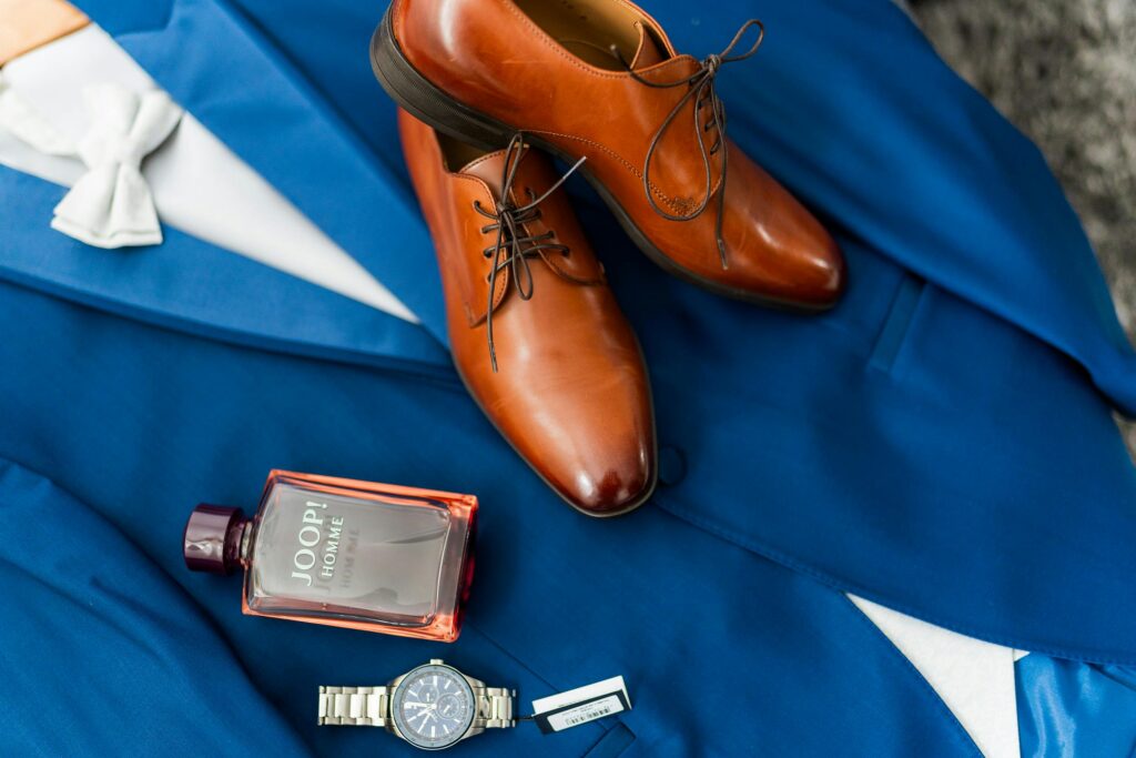 A stylish flat lay of blue suit with brown leather shoes, wristwatch, and perfume for a groom's special day.